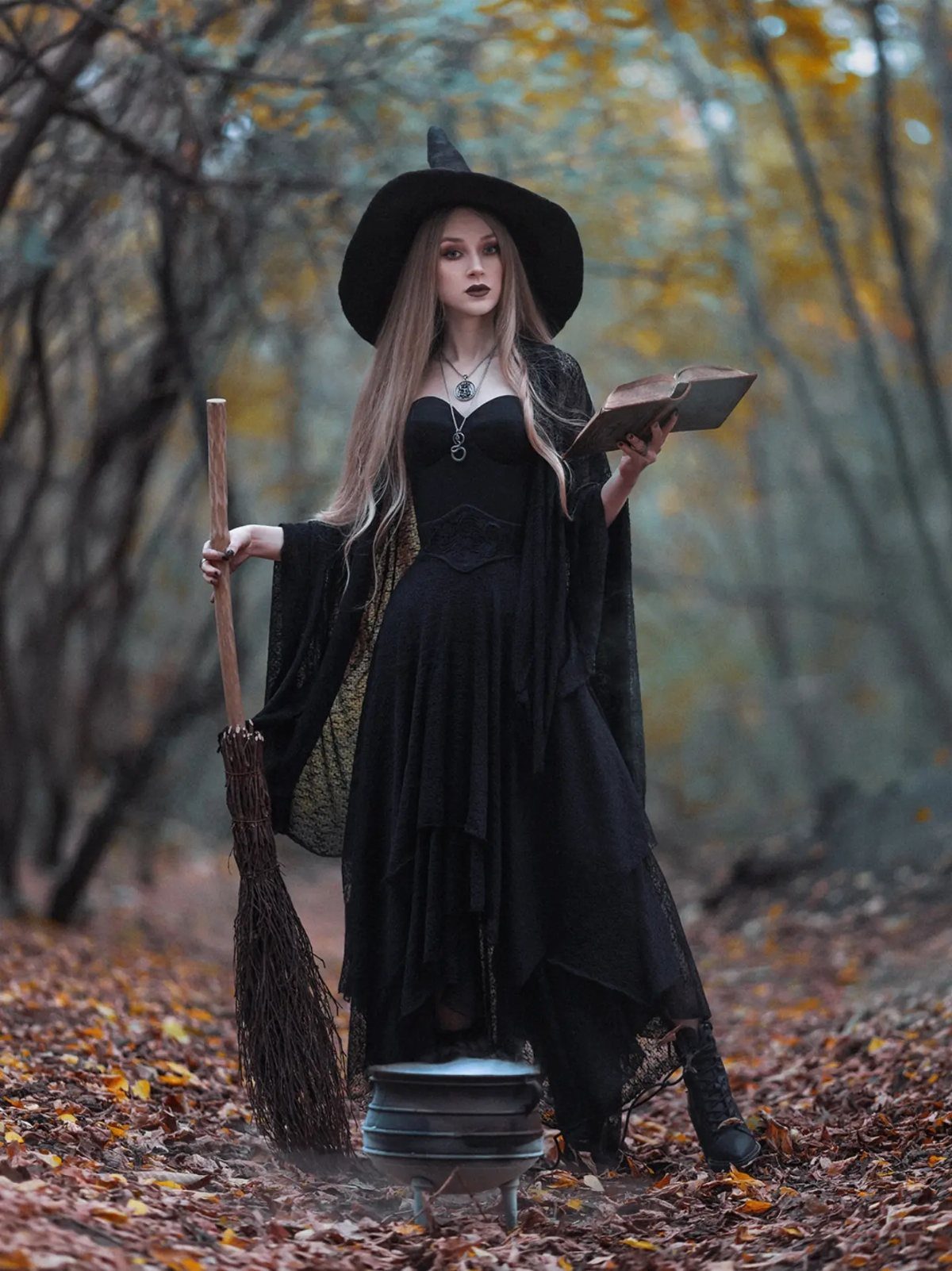 Woman in a black witch costume holding a broom and book in autumn woods