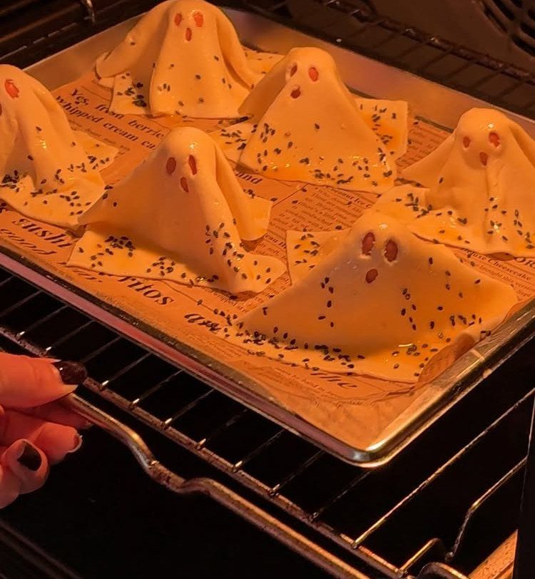 Ghost-shaped pastries with red eyes baking on a tray inside an oven.