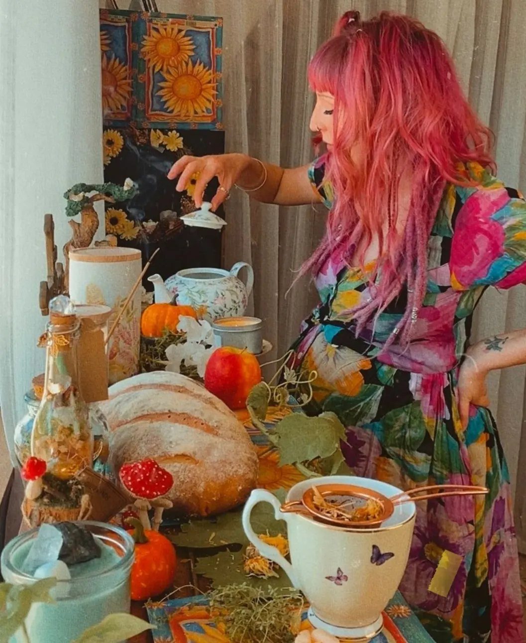 Woman with pink hair preparing tea at an earthy, bohemian style table setup.