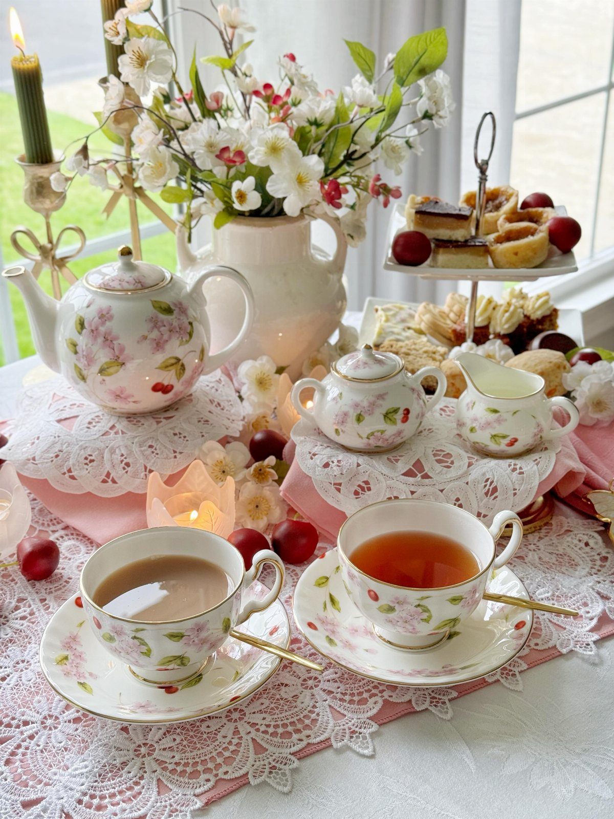 Floral teacups and teapot arranged with cakes and white blossoms on lace tablecloth