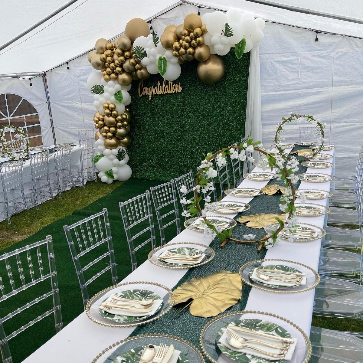 Party table with gold plates and a balloon garland against a green hedge backdrop