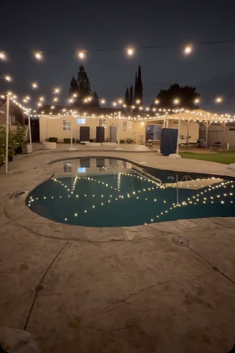 Warm white string lights draped over a backyard swimming pool and patio at night.