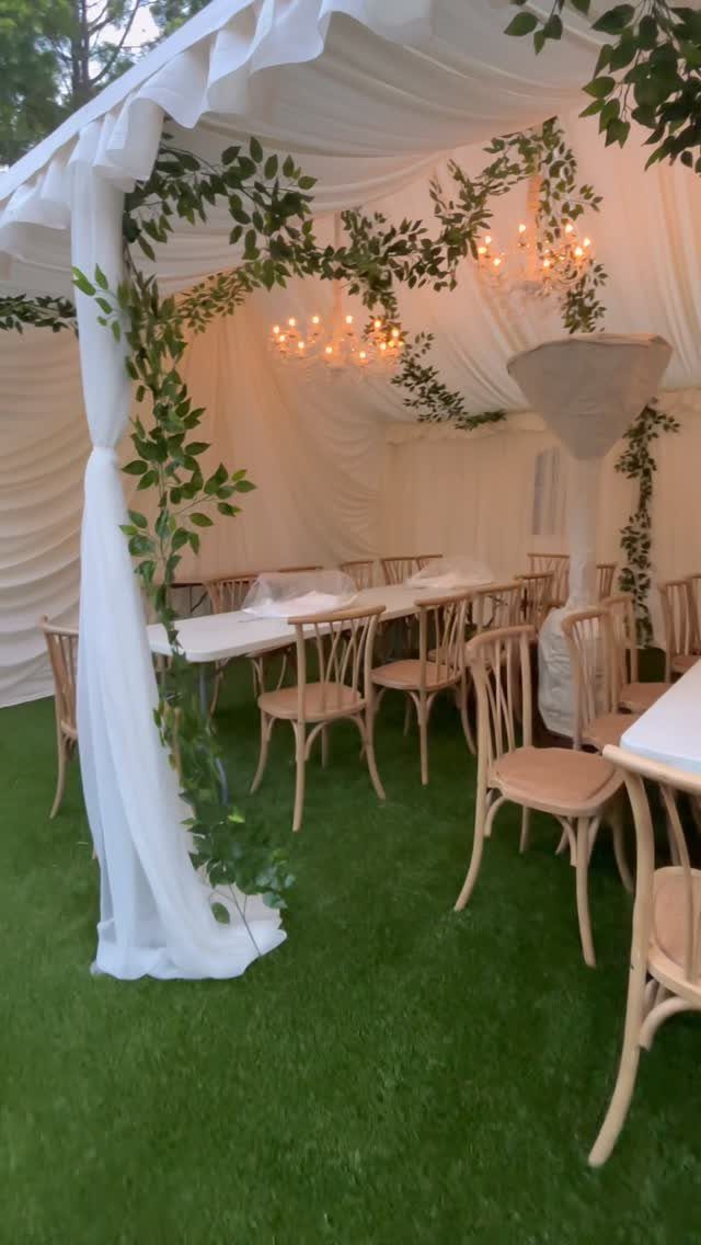 White canopy tent with a long dining table, wooden chairs, and hanging chandeliers.