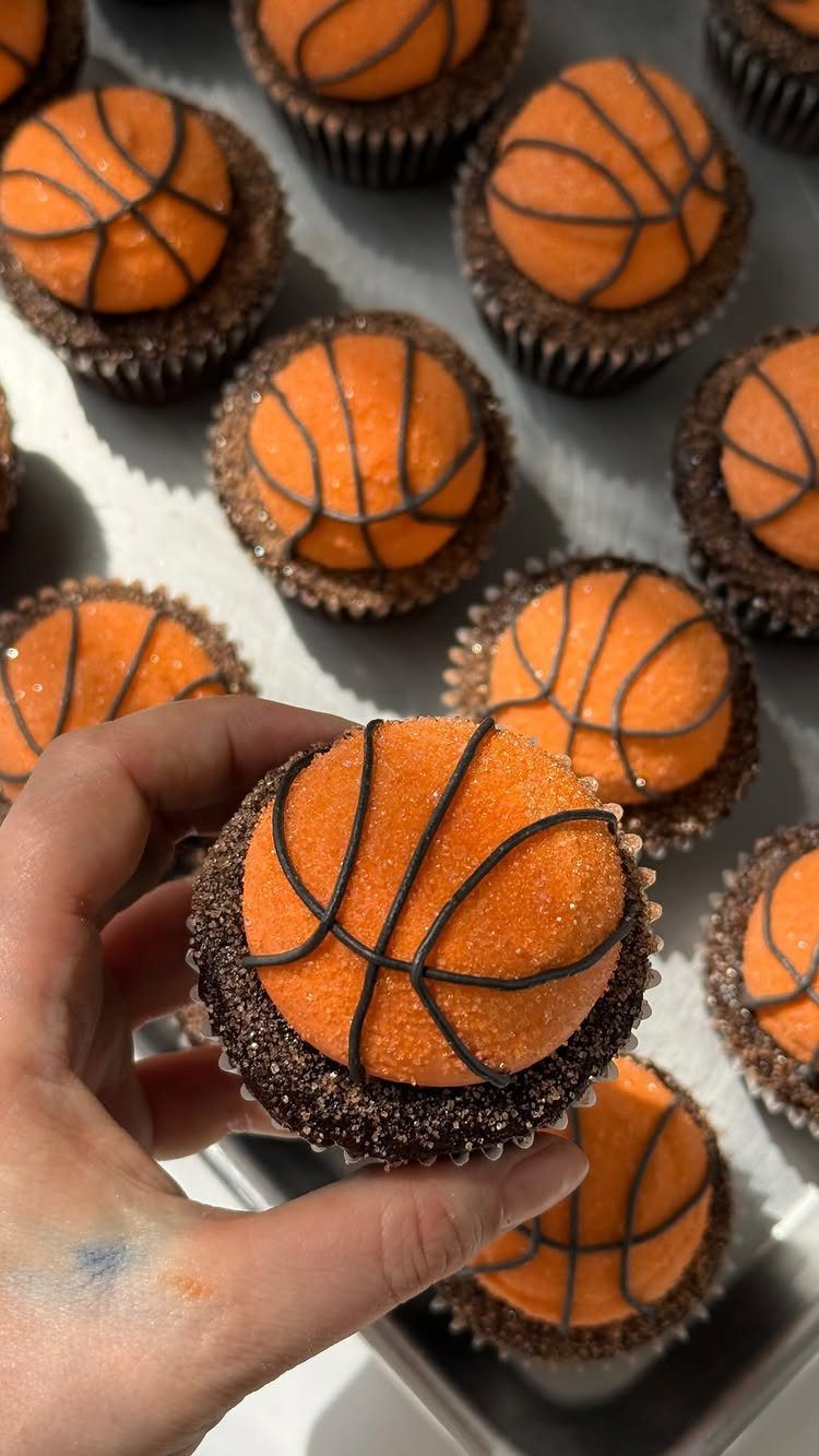 Chocolate cupcakes topped with orange basketball decorations held by a person's hand.