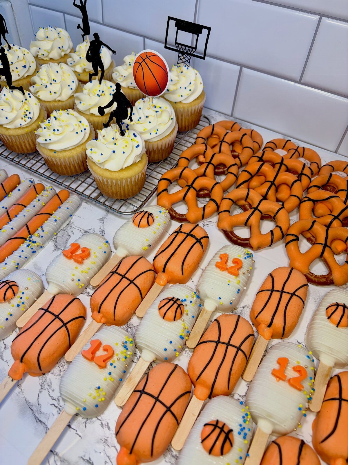 Basketball-themed treats including decorated popsicles, chocolate pretzels, and cupcakes on a marble surface.