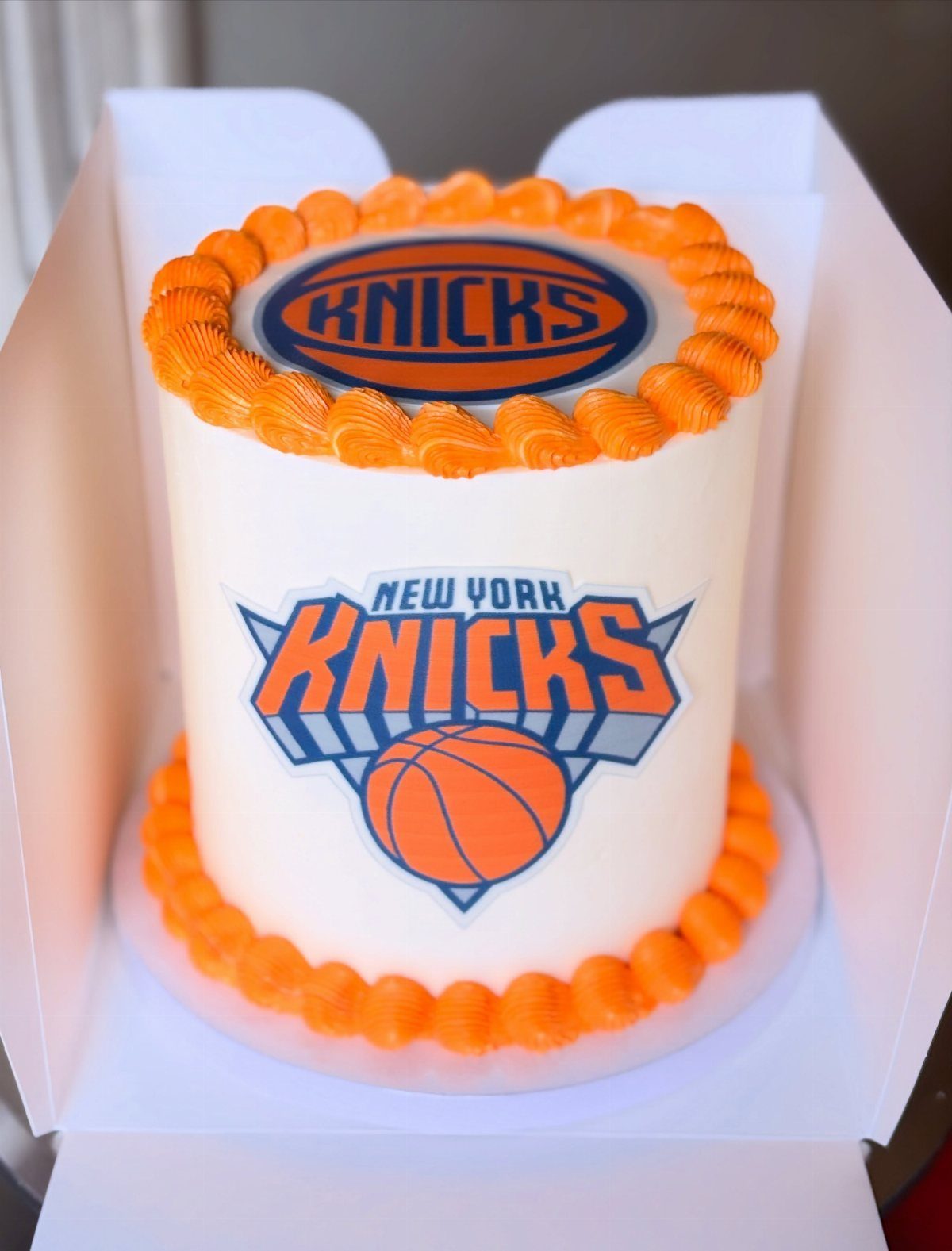 White cylindrical cake with orange frosting and New York Knicks basketball logos.