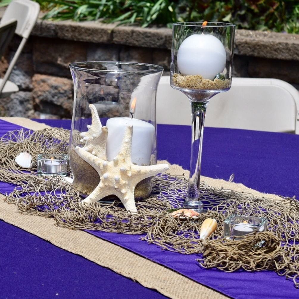 Beach themed table centerpieces with white candles, starfish, and fishing nets on purple cloth