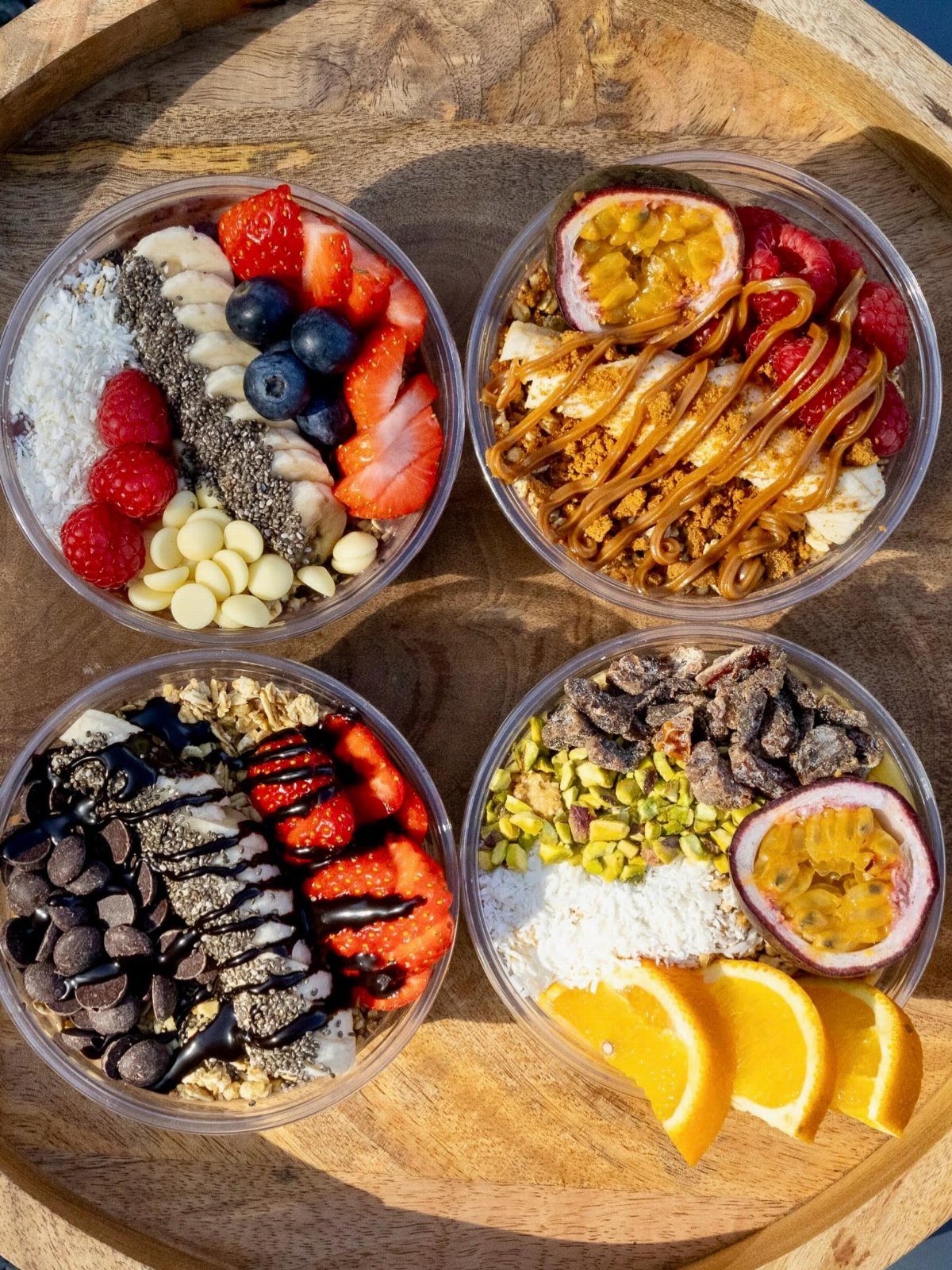 Four fruit smoothie bowls with fresh berries, passion fruit, and granola on wood.