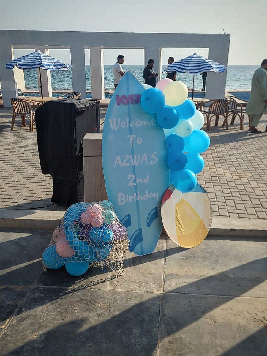 Beach party decorations with a surfboard cutout, balloons, and striped umbrellas by the sea.