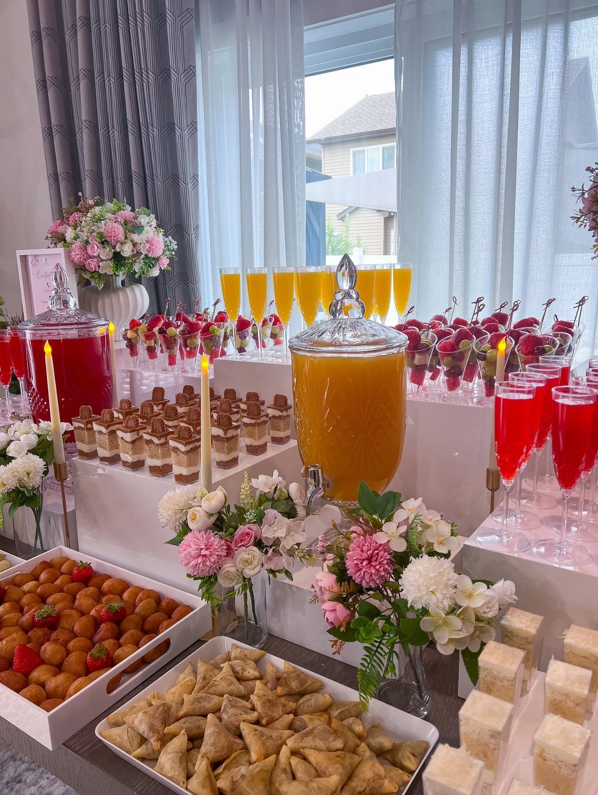 Buffet table with orange and red drinks, desserts, pastries, and pink floral arrangements.