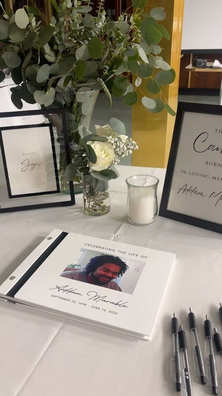 Memorial table with guest book, framed signs, white roses, and eucalyptus greenery.