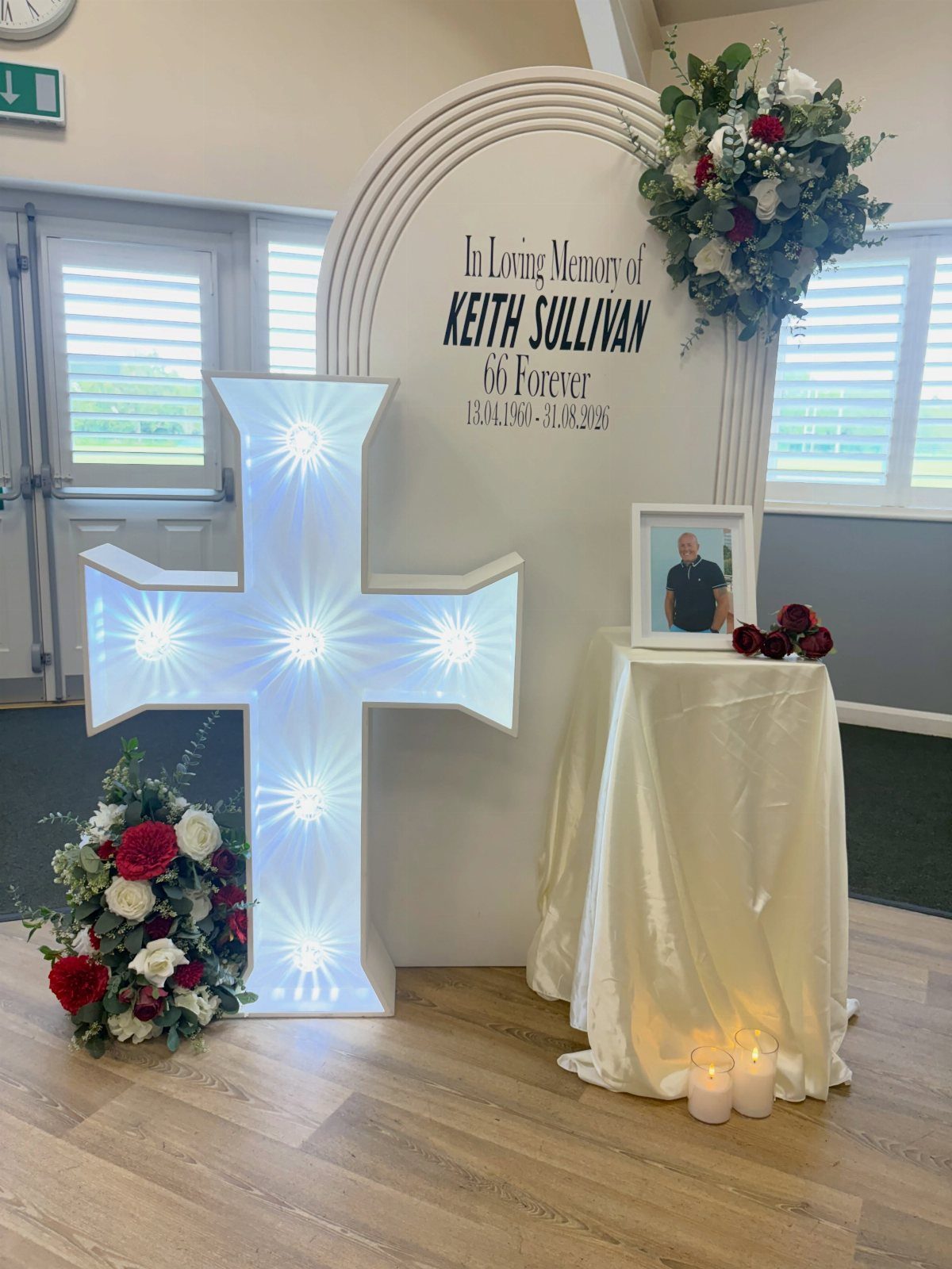 Memorial table with a framed portrait, glowing white cross, and floral arrangements.