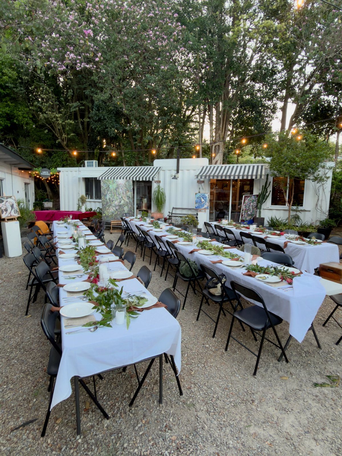 Long white dining tables with greenery centerpieces set outdoors under string lights.