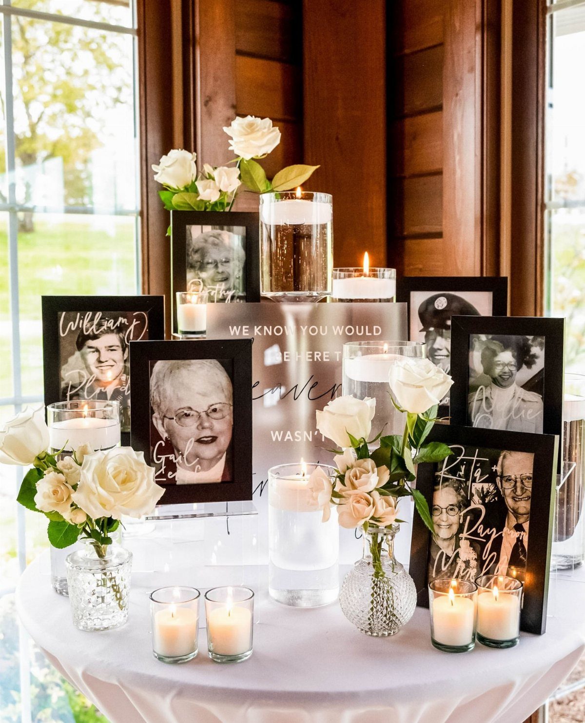 Memory table with framed portraits, white roses, and lit candles on white cloth.