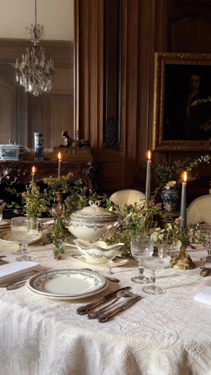 Elegant dining table with white lace cloth, lit candles, and greenery centerpieces.