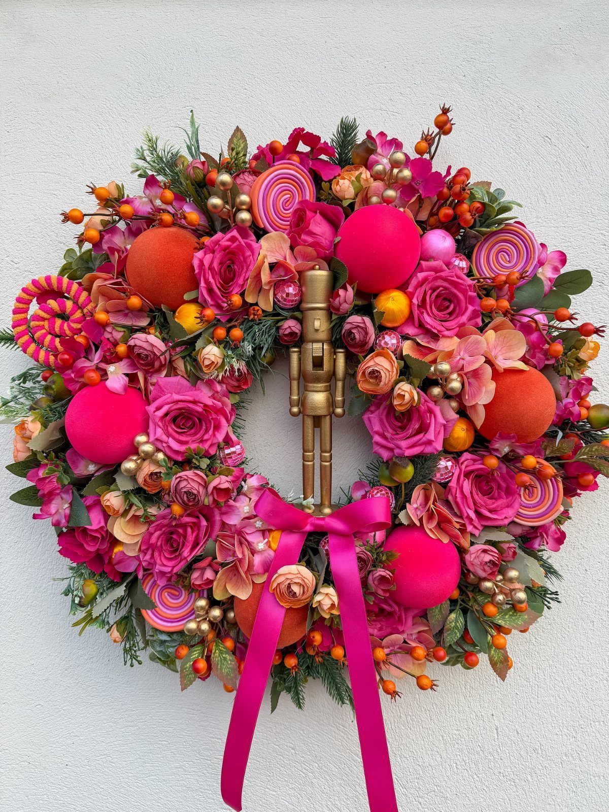 Pink and orange Christmas wreath with roses, a golden nutcracker, and pink ribbon.
