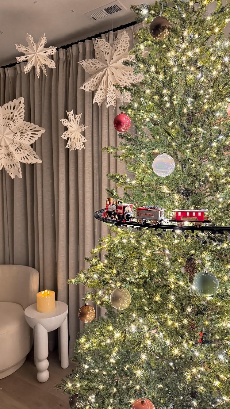 White oversized paper snowflakes hanging by a lit Christmas tree and grey curtains