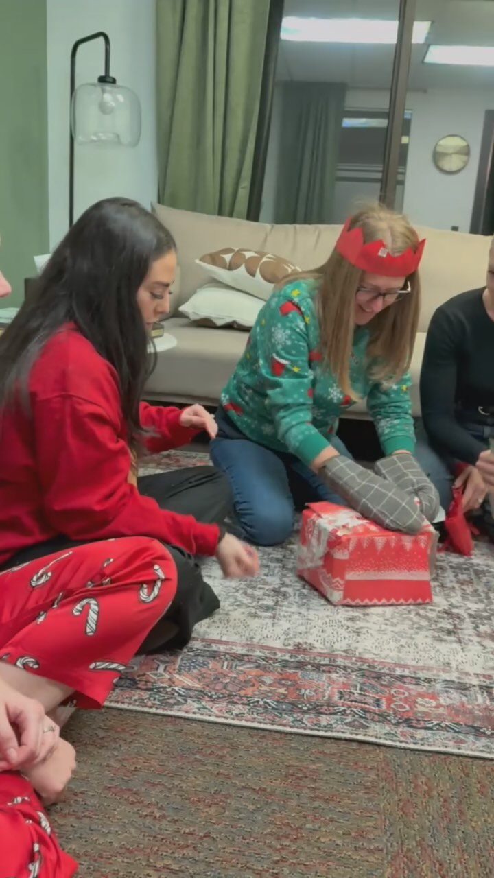 Women in Christmas pajamas playing a gift-opening game with oven mitts on a rug