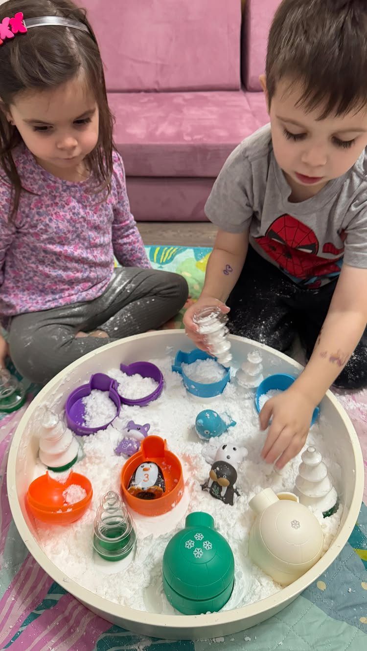 Two children playing with artificial snow and snowman molds in a white bin.
