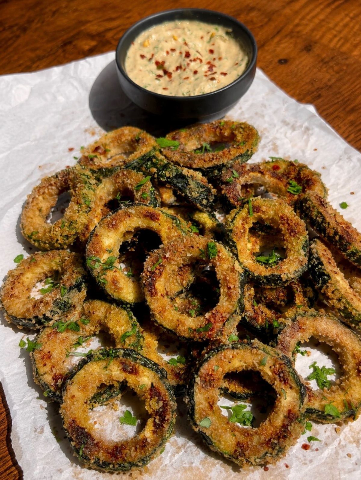 Breaded zucchini chips on parchment paper with a side of creamy dipping sauce.