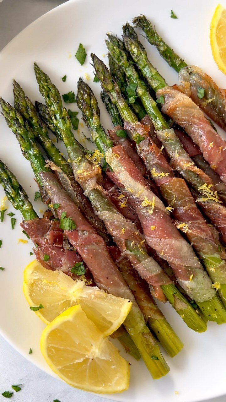Prosciutto-wrapped asparagus spears garnished with lemon zest and parsley on a white plate.