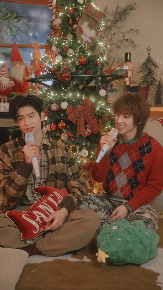 Two men singing into microphones while sitting by a decorated Christmas tree.