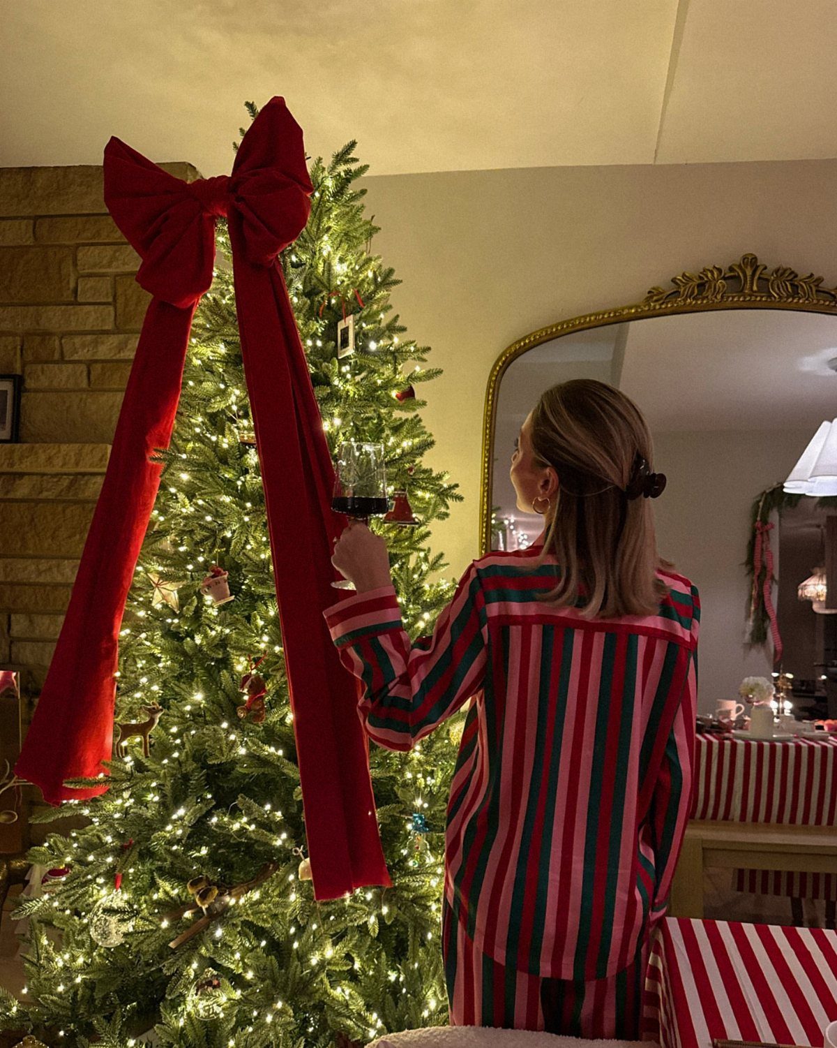 Woman in striped Christmas pajamas holding wine next to a decorated Christmas tree.