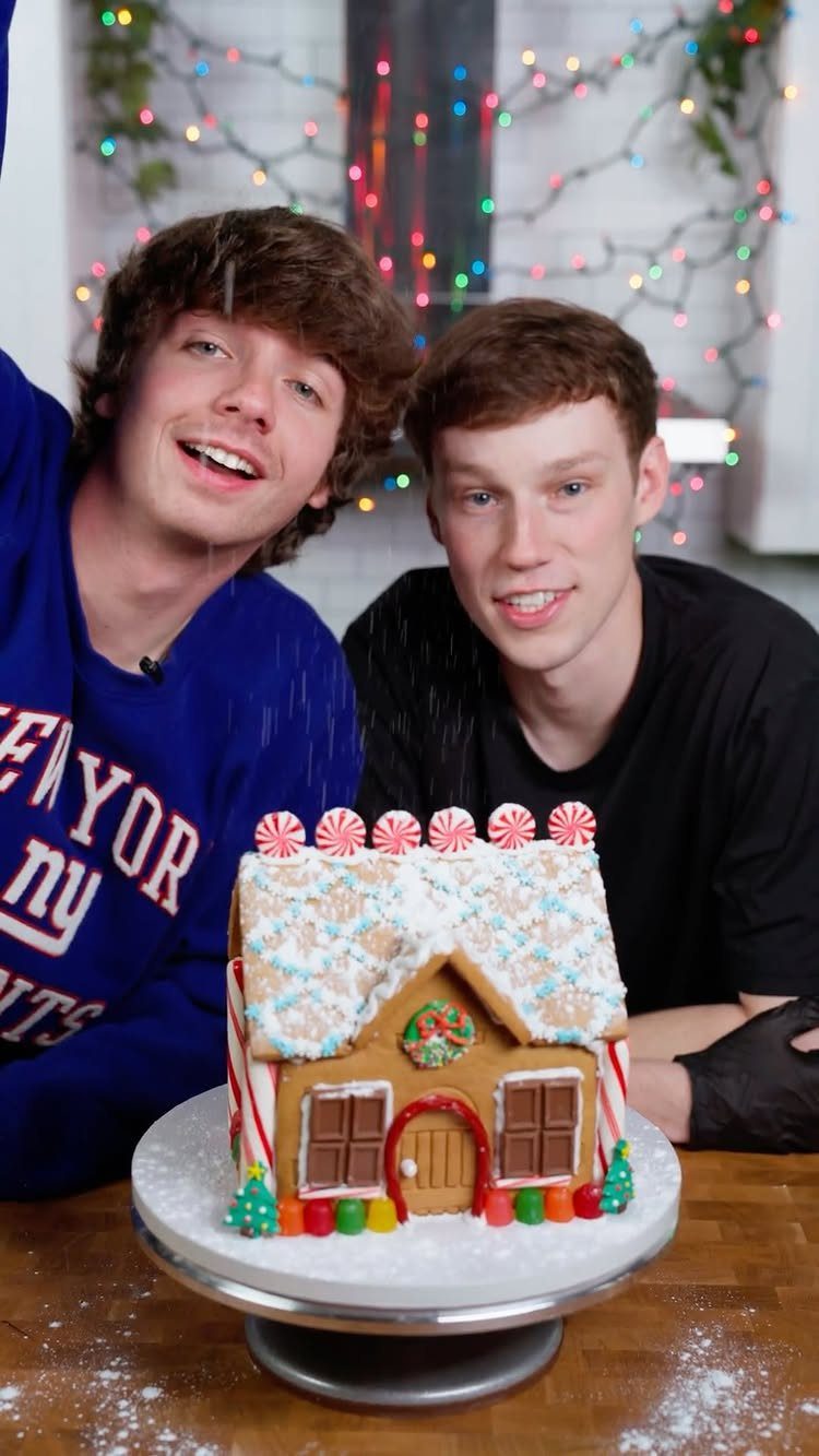 Two men smiling behind a decorated gingerbread house with peppermint and gumdrops.