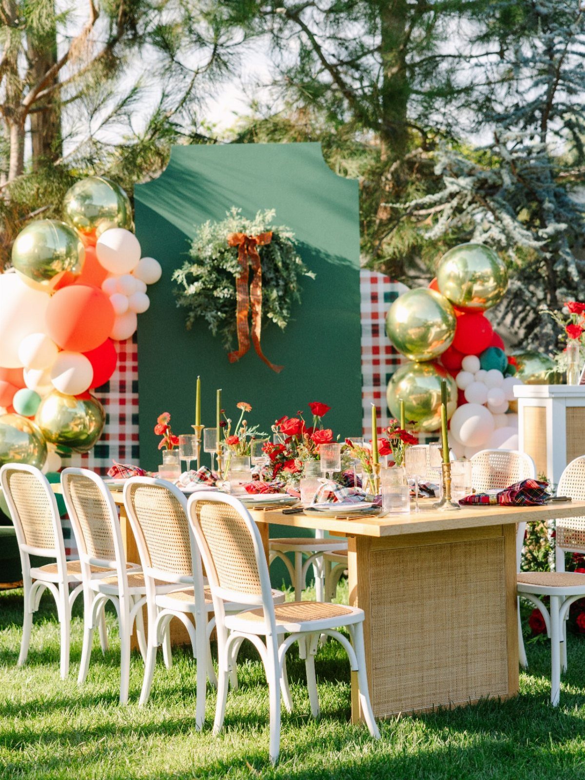 Outdoor Christmas party table setting with red flowers, green backdrop, and festive balloons.