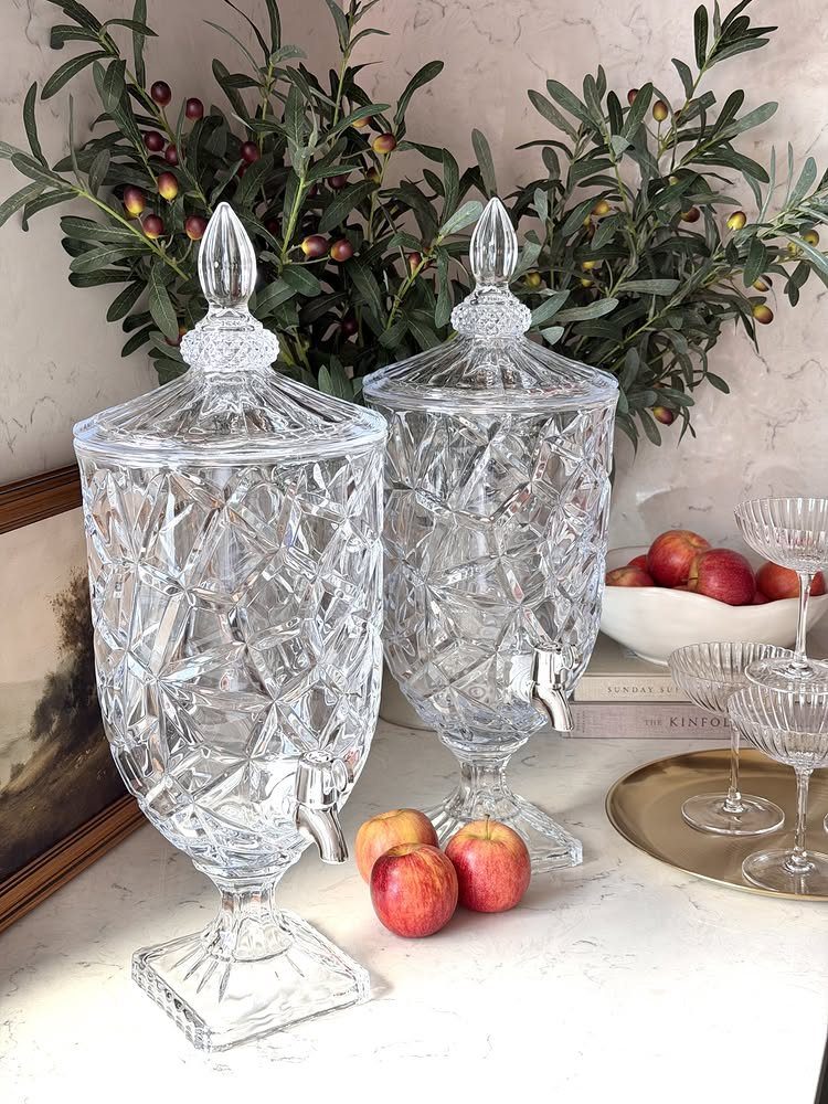 Two crystal glass beverage dispensers with spigots on a marble table with apples.