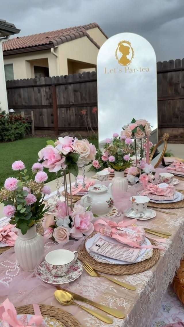Pink tea party table setting with floral arrangements, teacups, and gold cutlery outdoors.