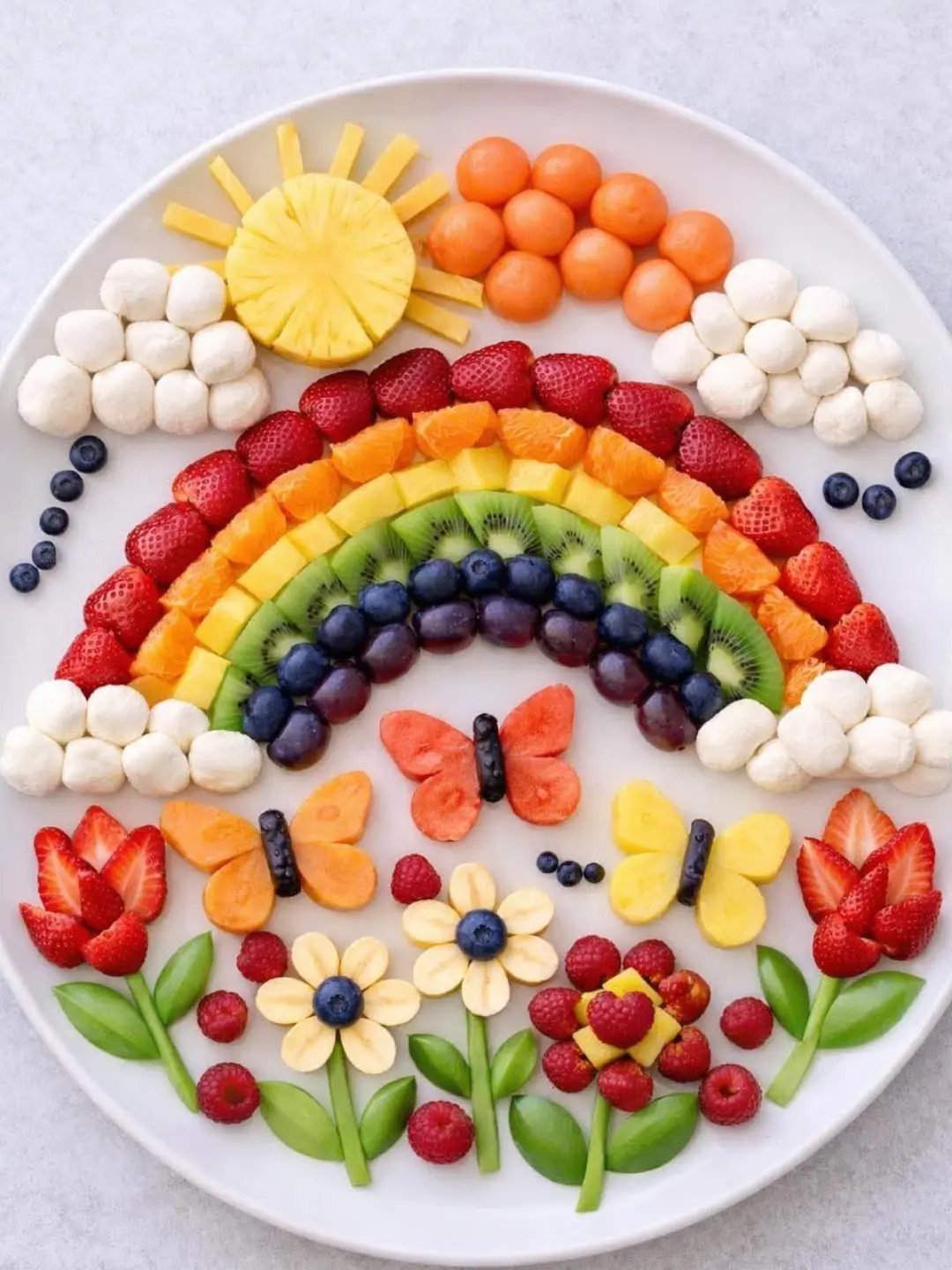 Fruit platter arranged as a rainbow landscape with a sun, clouds, and flowers.