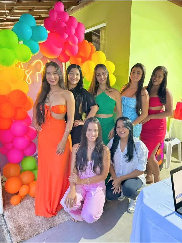 Six women in colorful solid outfits posing before a vibrant rainbow balloon arch.