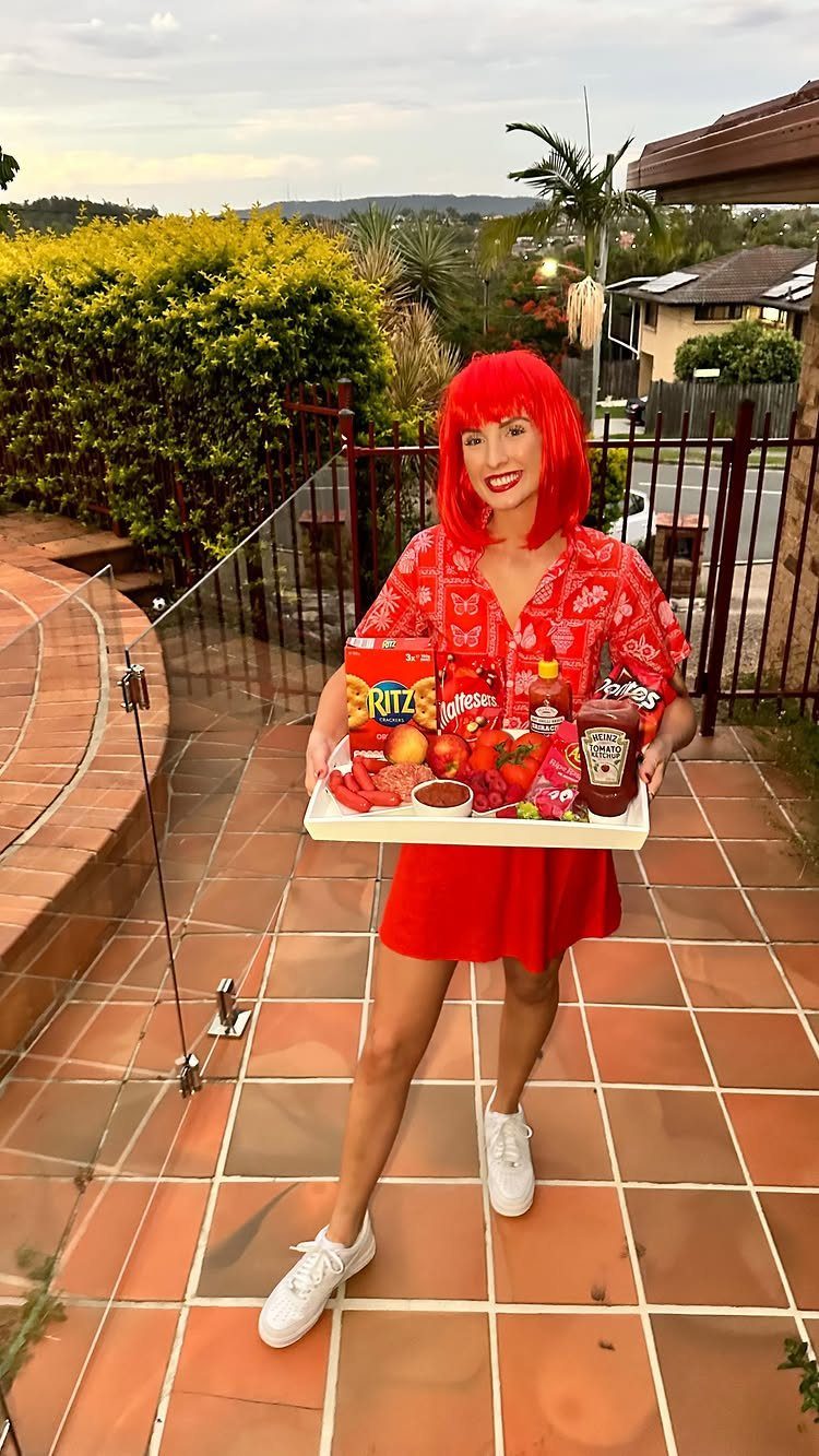 Woman in red dress and wig holding a tray of red snacks.