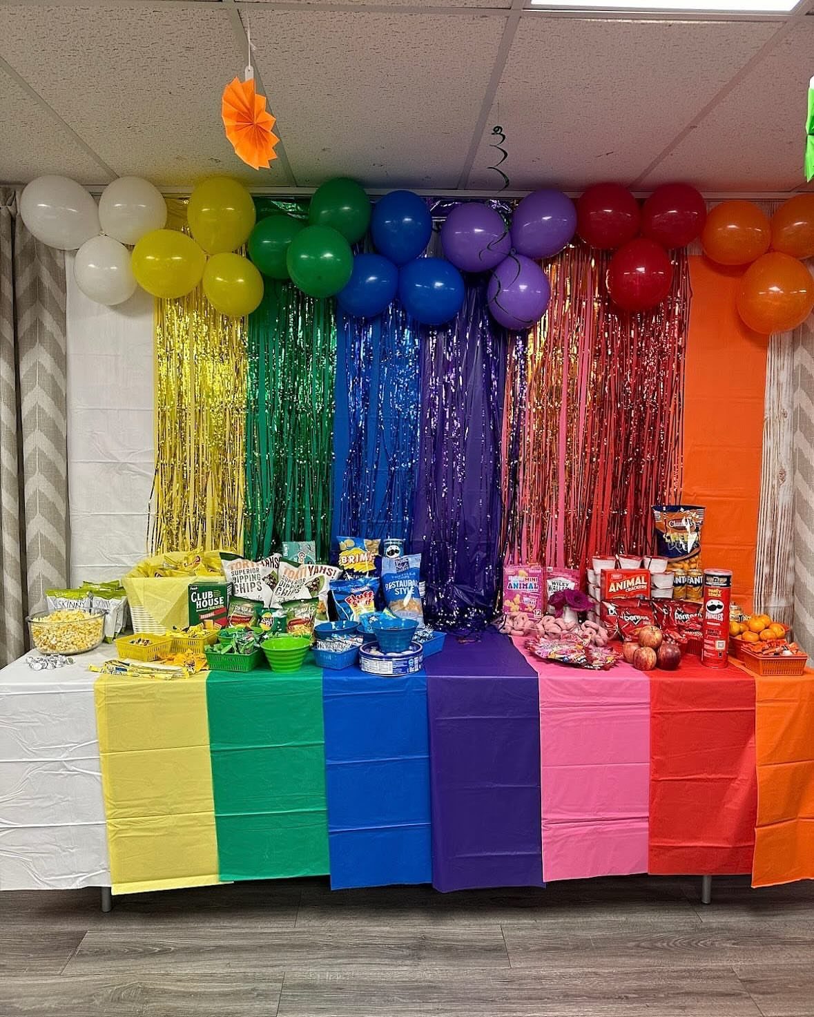 Rainbow colored party table with matching snacks, tinsel curtains, and balloons in stripes.