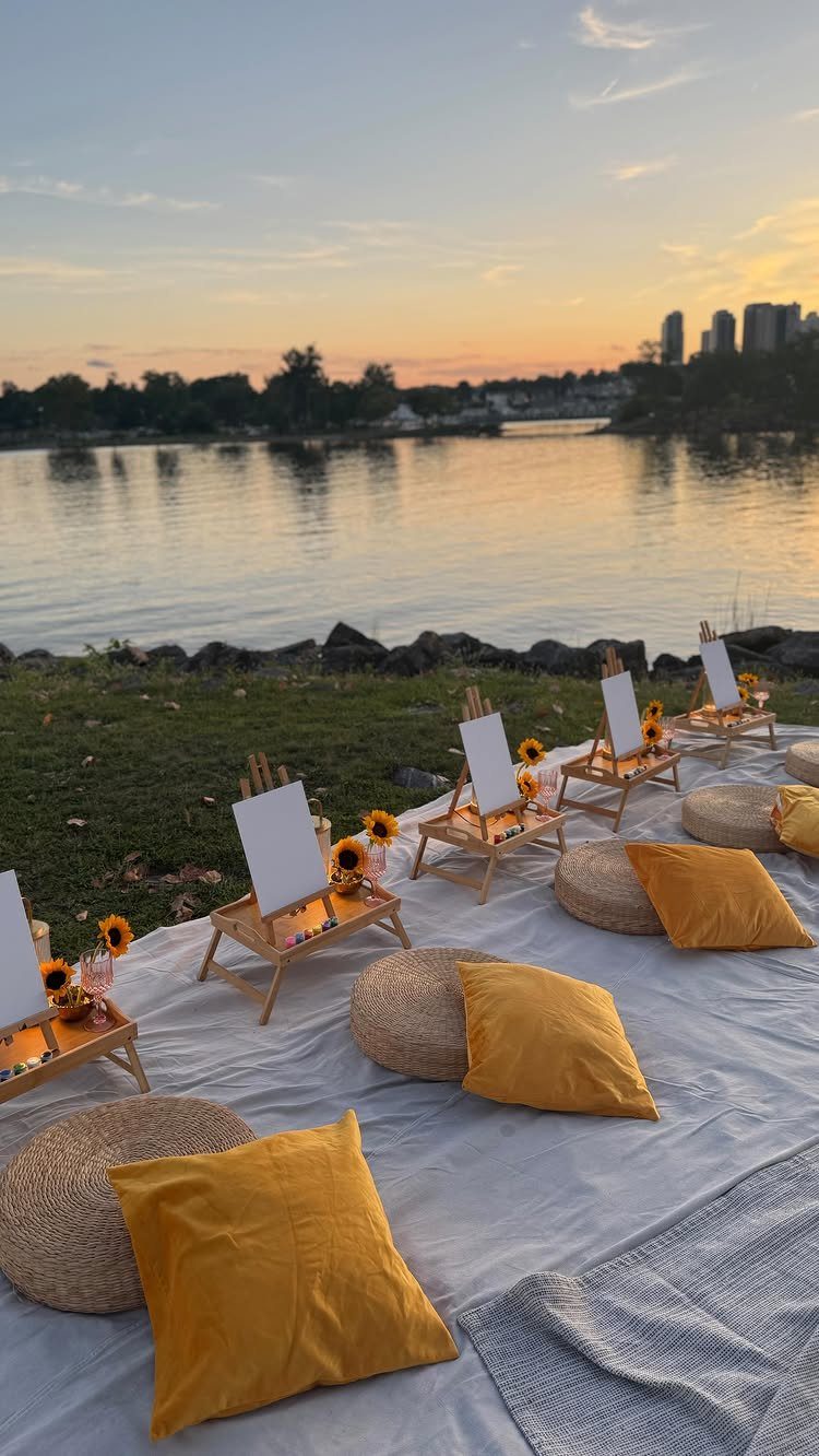 Yellow themed picnic setup with pillows and sunflowers by a lake at sunset