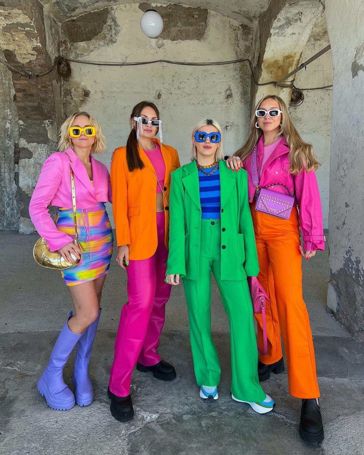 Four women wearing bright neon blazers and colorful outfits posing in a concrete room.