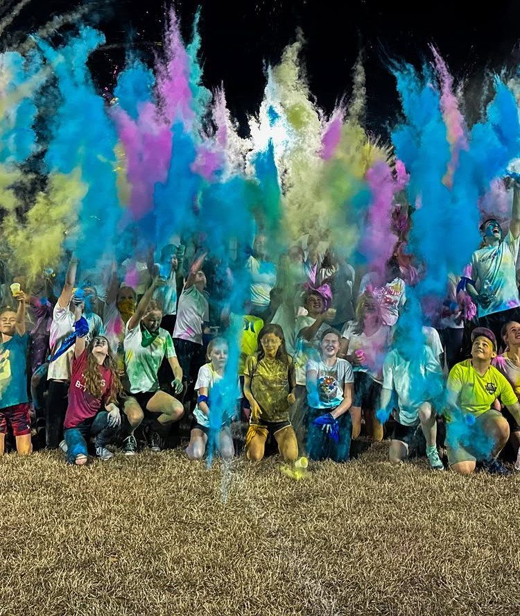 People throwing vibrant blue, pink, and yellow color powders in a grassy field.