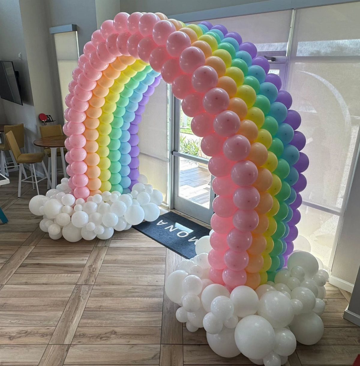 Rainbow balloon arch with white balloon clouds on a wooden floor indoors.