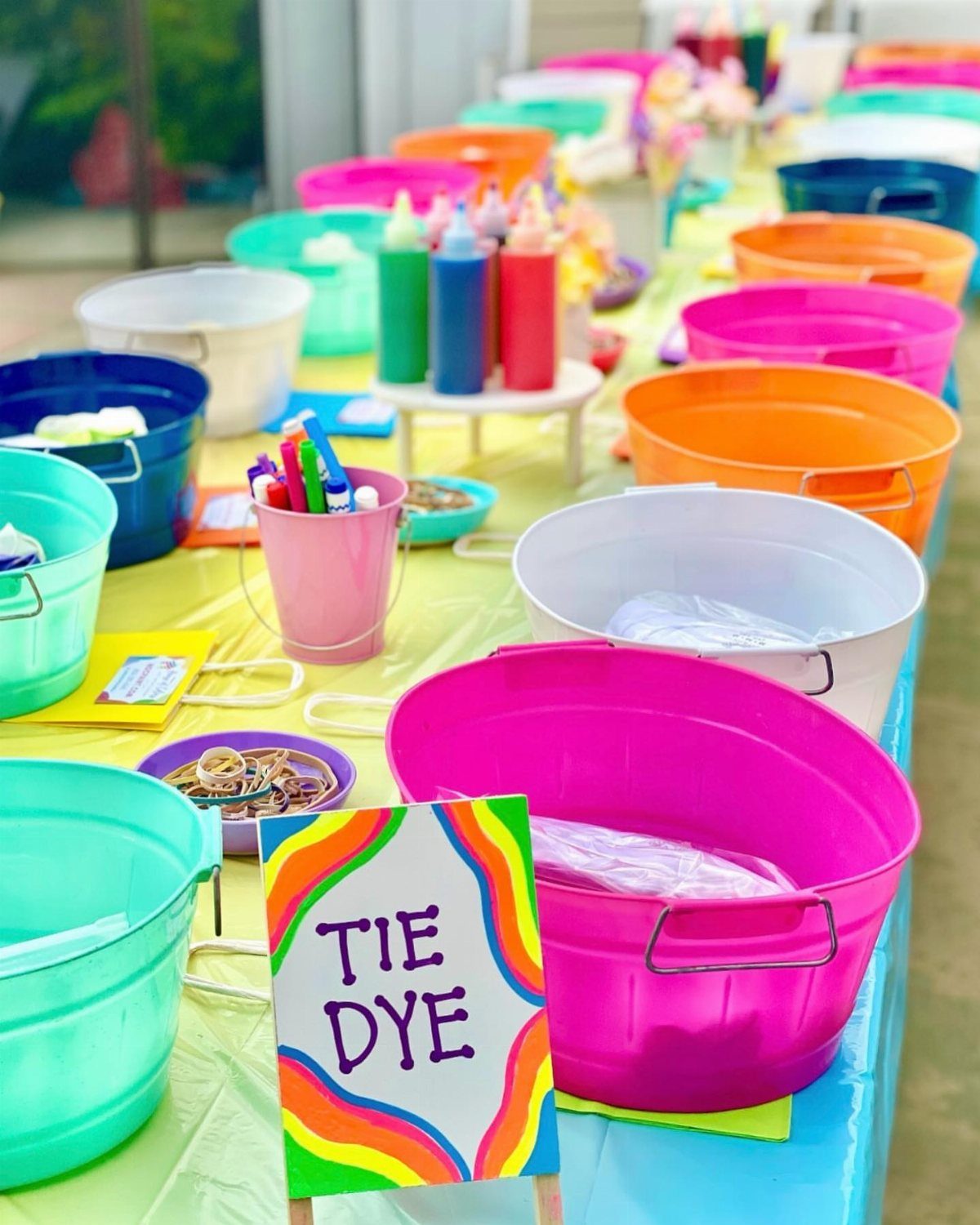 Tie-dye station with colorful plastic tubs and dye bottles on a yellow table.