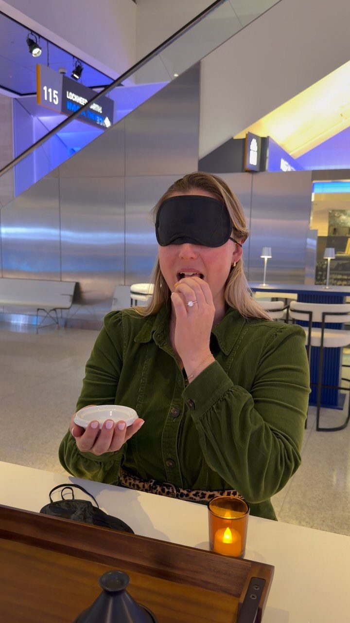 Blindfolded woman in green jacket tasting food in a modern indoor space.