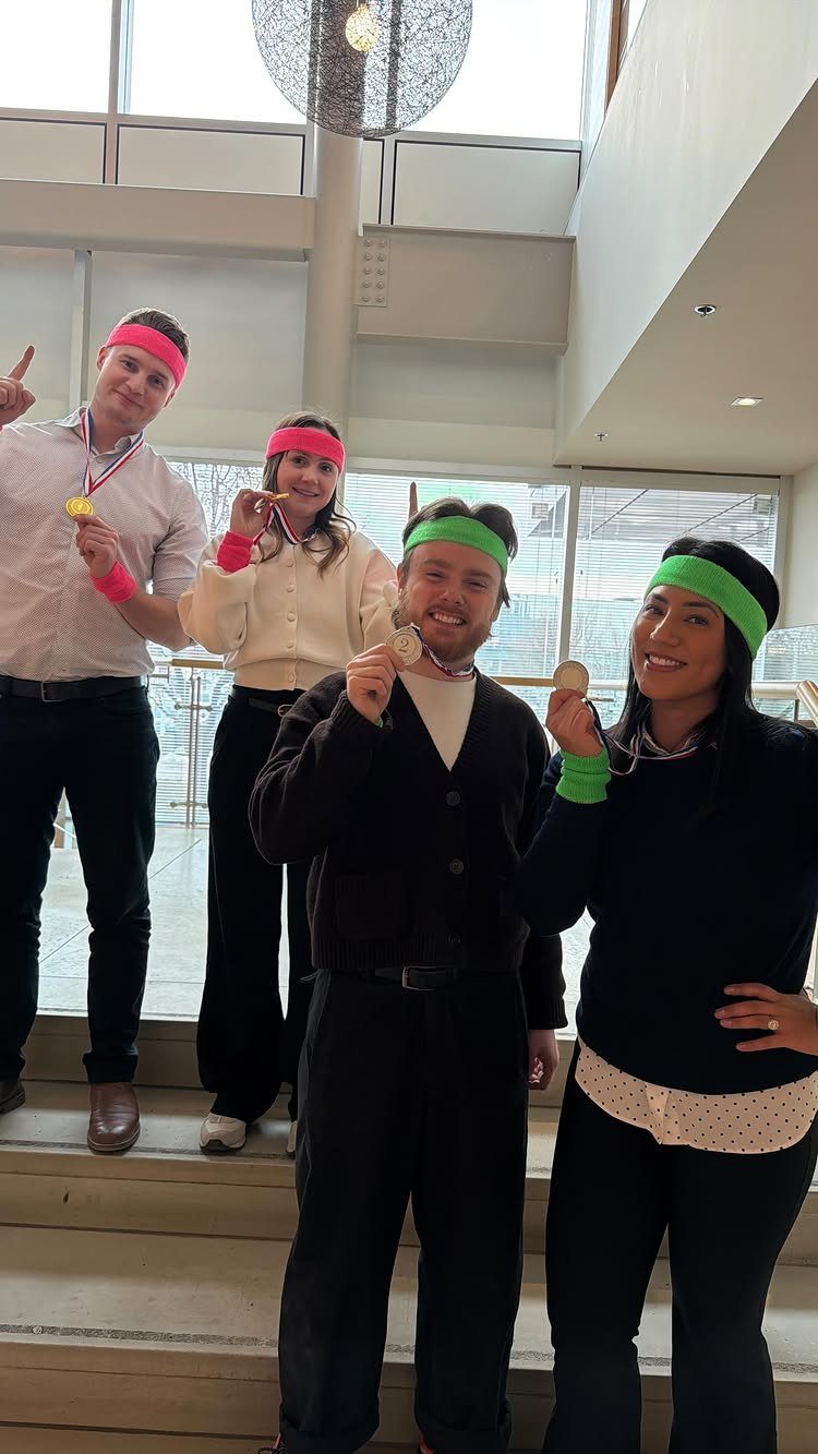Four smiling coworkers wearing colorful sweatbands and gold medals in a modern office.