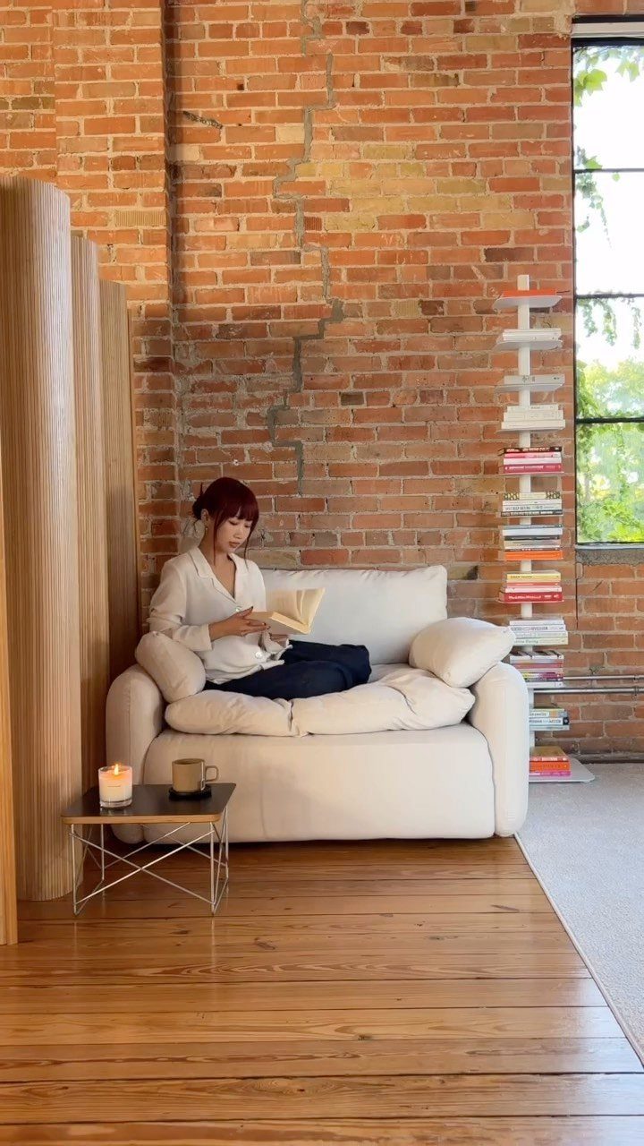 Woman reading a book on a white cozy chair against a brick wall.