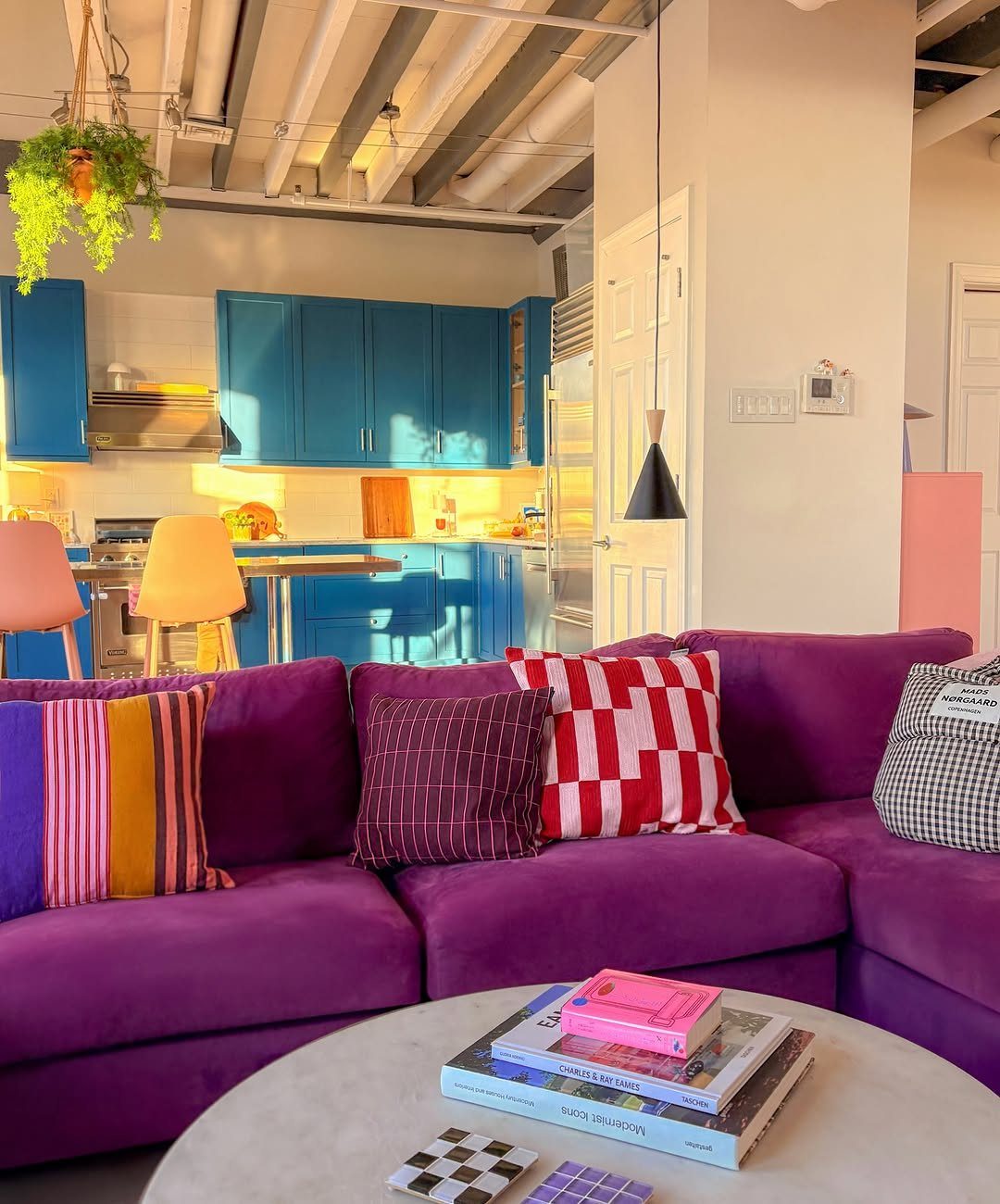 Purple sectional sofa with colorful pillows and blue kitchen cabinets in a bright room