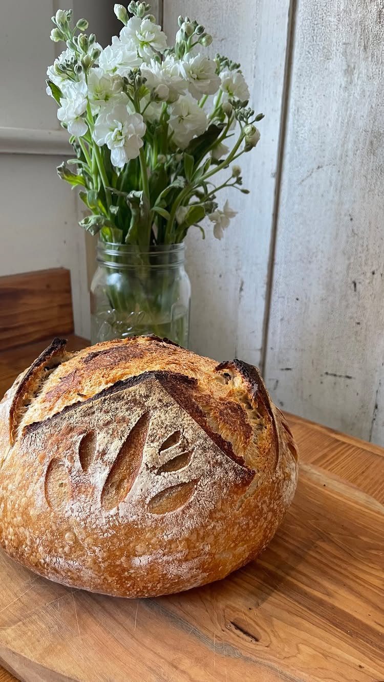 Artisan sourdough bread loaf with leaf scoring on a wooden board with white flowers.