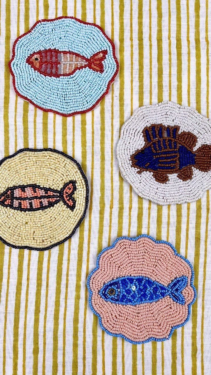 Four beaded shell-shaped coasters with colorful fish designs on a striped yellow cloth.
