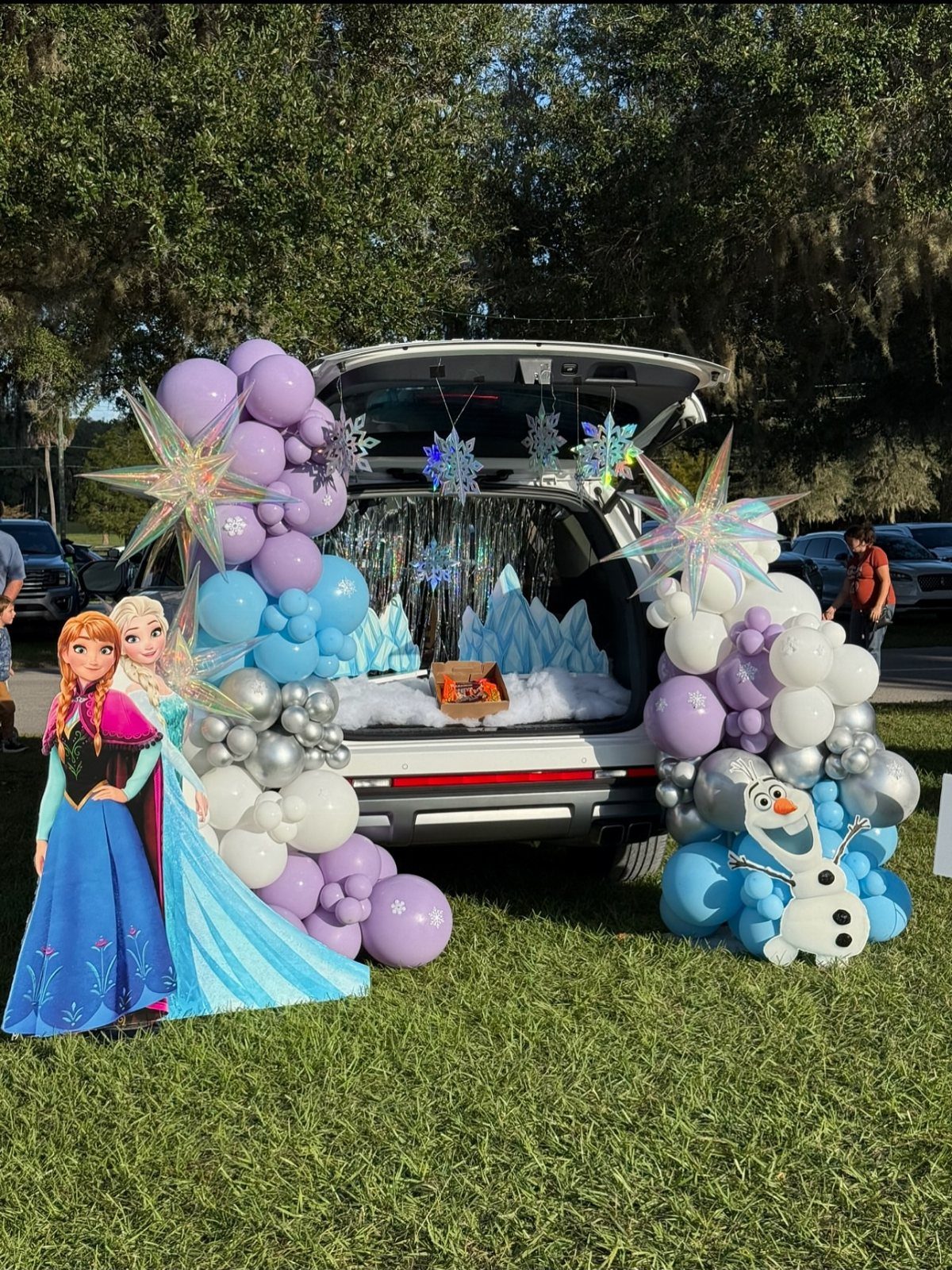 Frozen themed car trunk decoration with balloons, snowflakes, and character cutouts on grass.