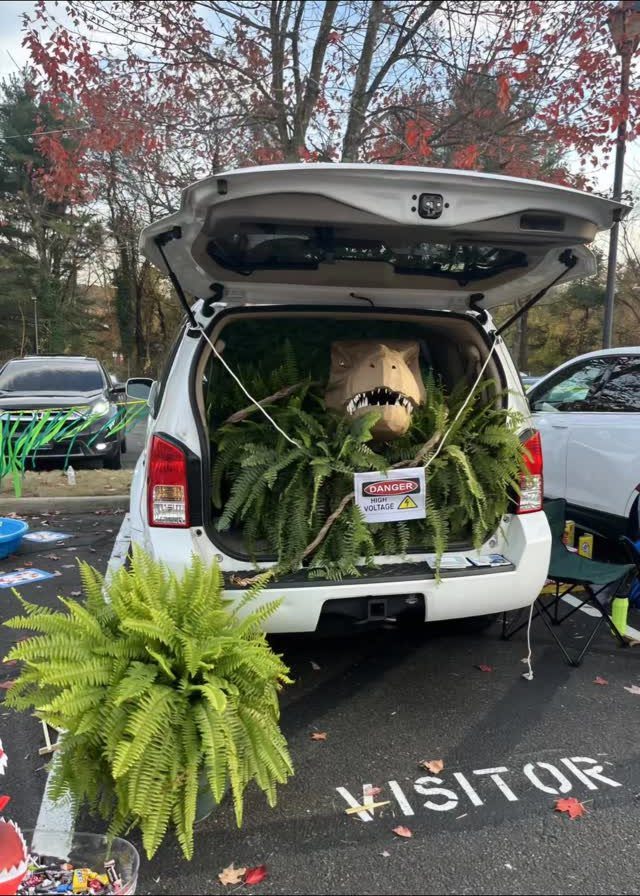 Dinosaur head emerging from green ferns in the trunk of a white SUV.