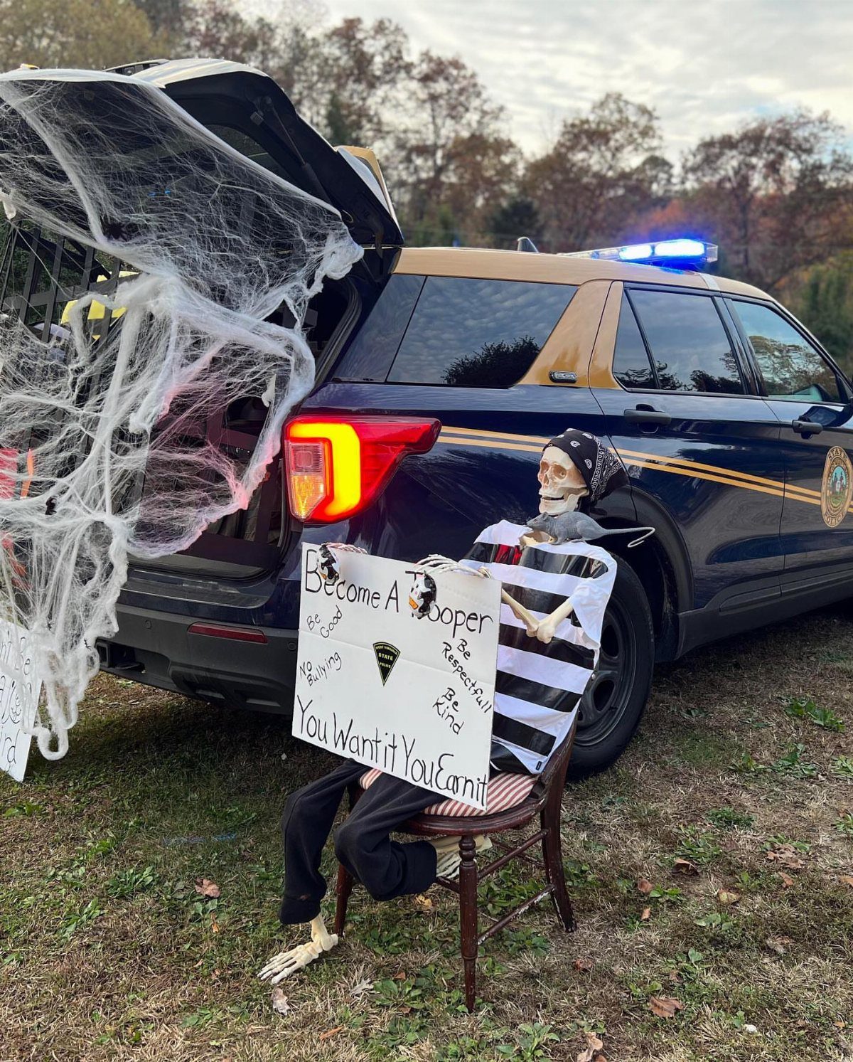 Skeleton in a striped shirt sitting by a police car with spider webs.
