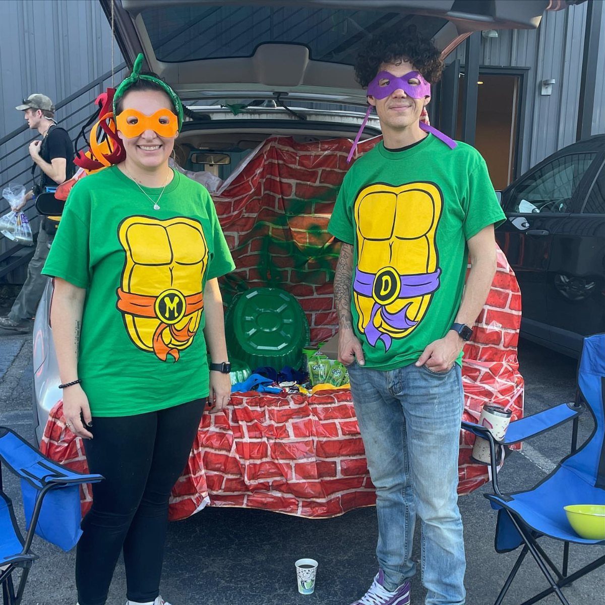 Two people in Teenage Mutant Ninja Turtles costumes posing by a decorated car trunk.